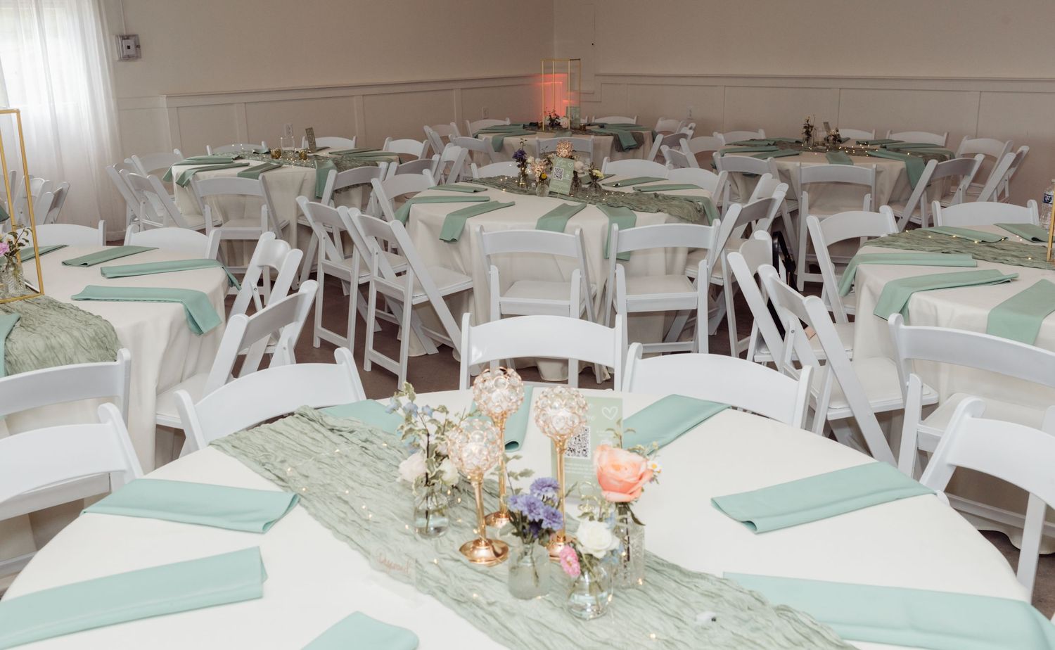Wedding reception hall with round tables featuring sage green napkins, textured runners, and floral centerpieces.