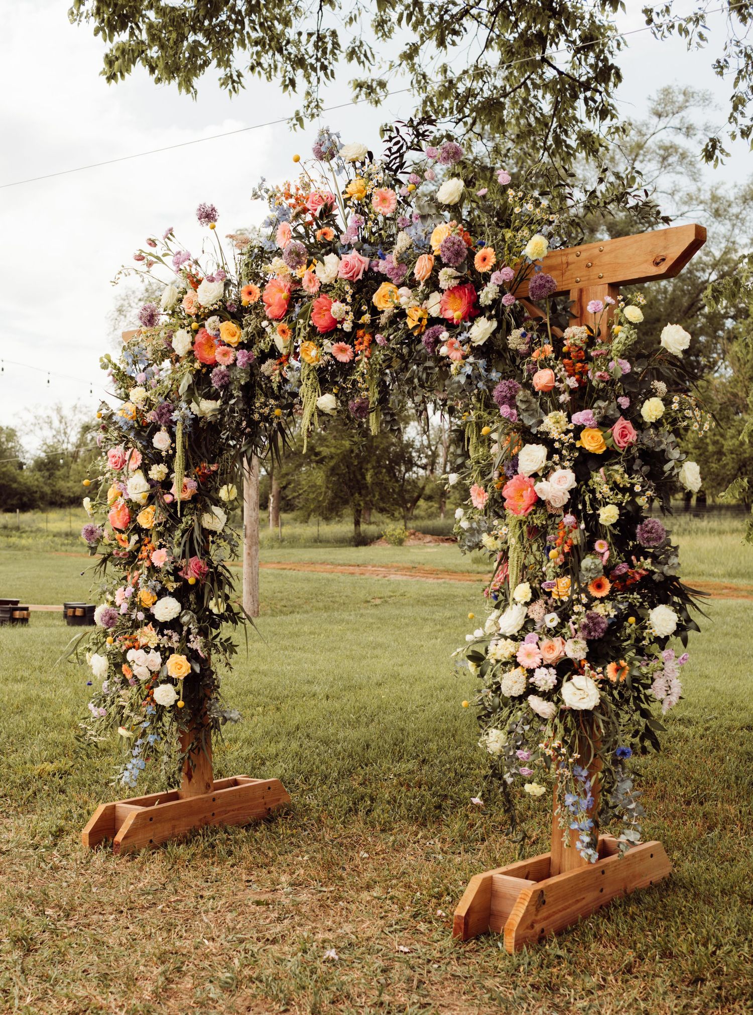 Lush outdoor wedding arch adorned with colorful flowers including roses, ranunculus, and wildflowers on green lawn.