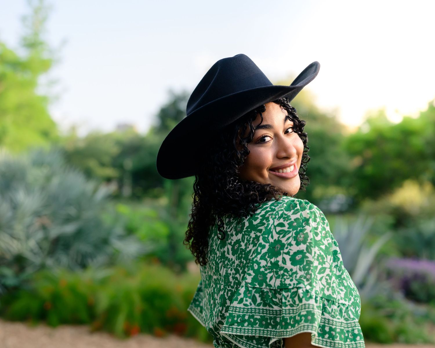 A smiling person wearing a black cowboy hat and green floral top poses against a blurred garden background.