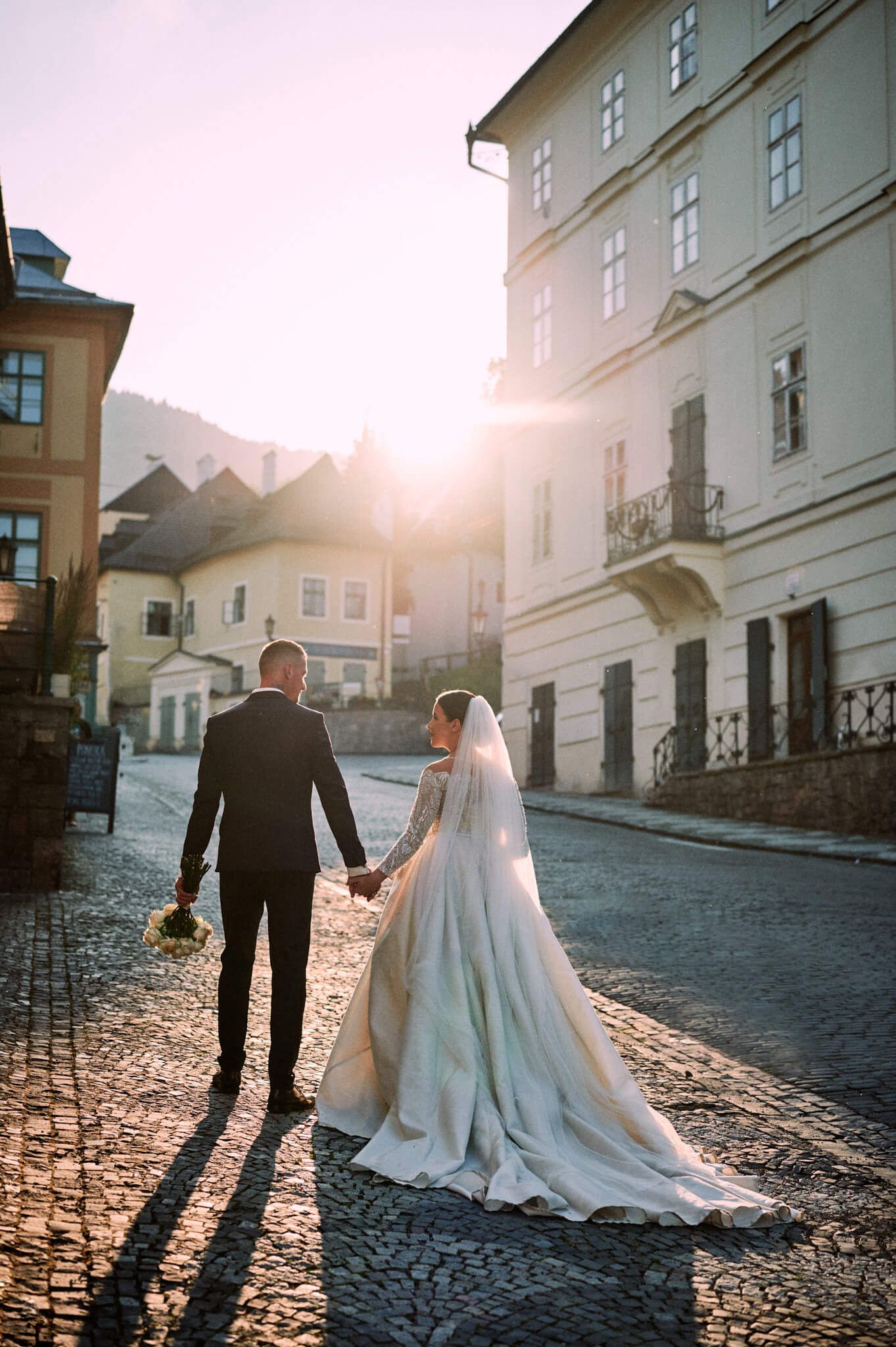 Couple walks hand in hand down cobblestone street during golden hour in historic city setting.