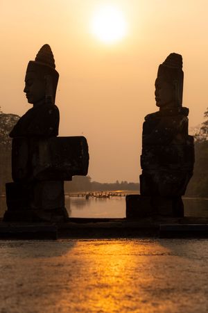 Stone statues silhouetted against golden sunset reflecting on water at Angkor Wat.