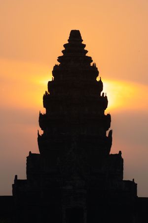 Dramatic silhouette of an ancient Khmer temple spire against an orange sunset sky.