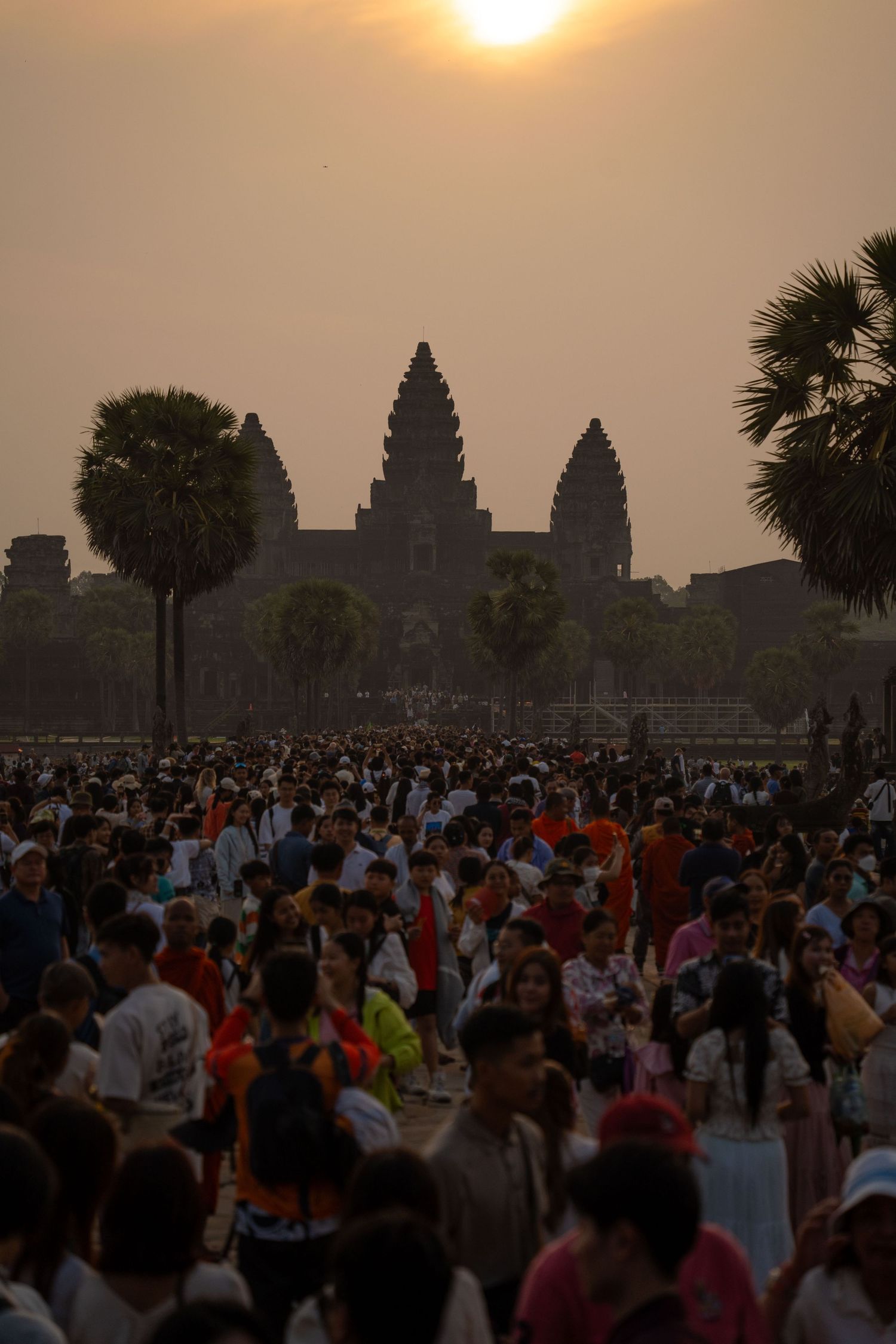 Crowds gather at Angkor Wat temple complex during sunset with palm trees framing the iconic spires.