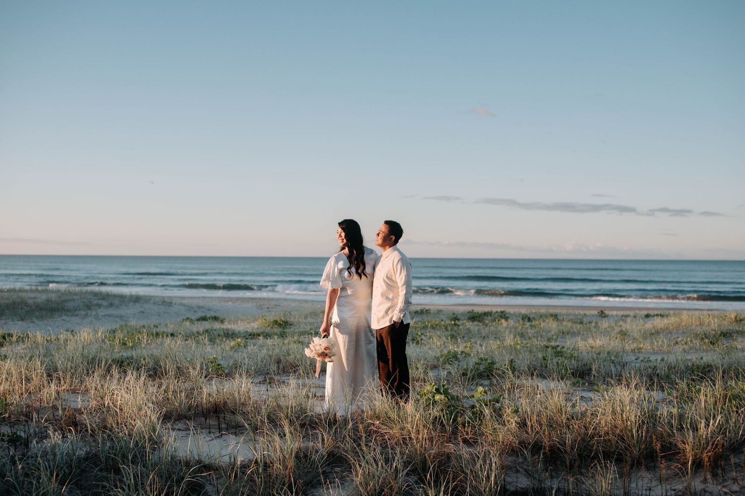 A couple stands together on a coastal hilltop during golden hour overlooking the ocean and grassy dunes.