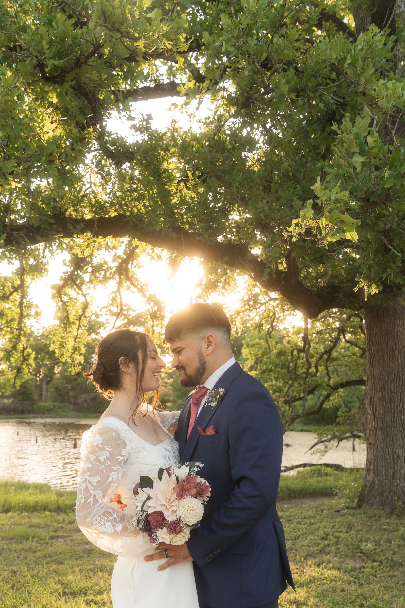 Two newlyweds share an intimate moment at sunset under a large oak tree by a lake, with golden sunlight filtering through.