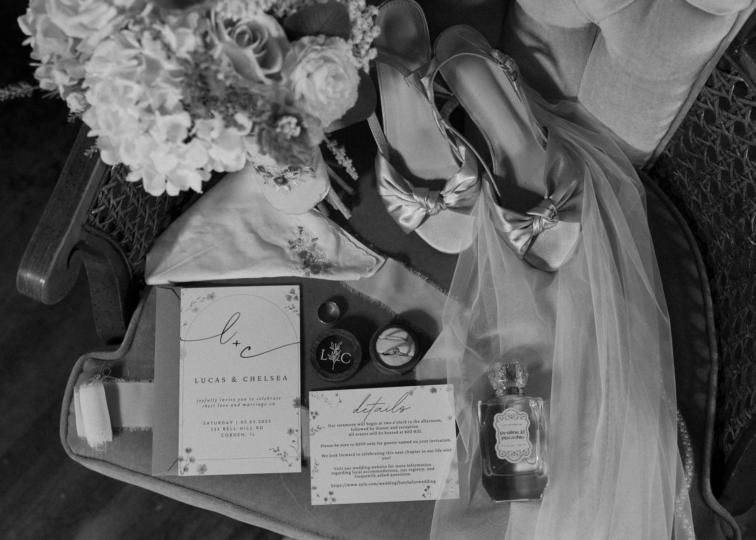 Wedding details arranged on dark surface include invitations, high heel shoes, perfume bottle, veil and floral bouquet in black and white.