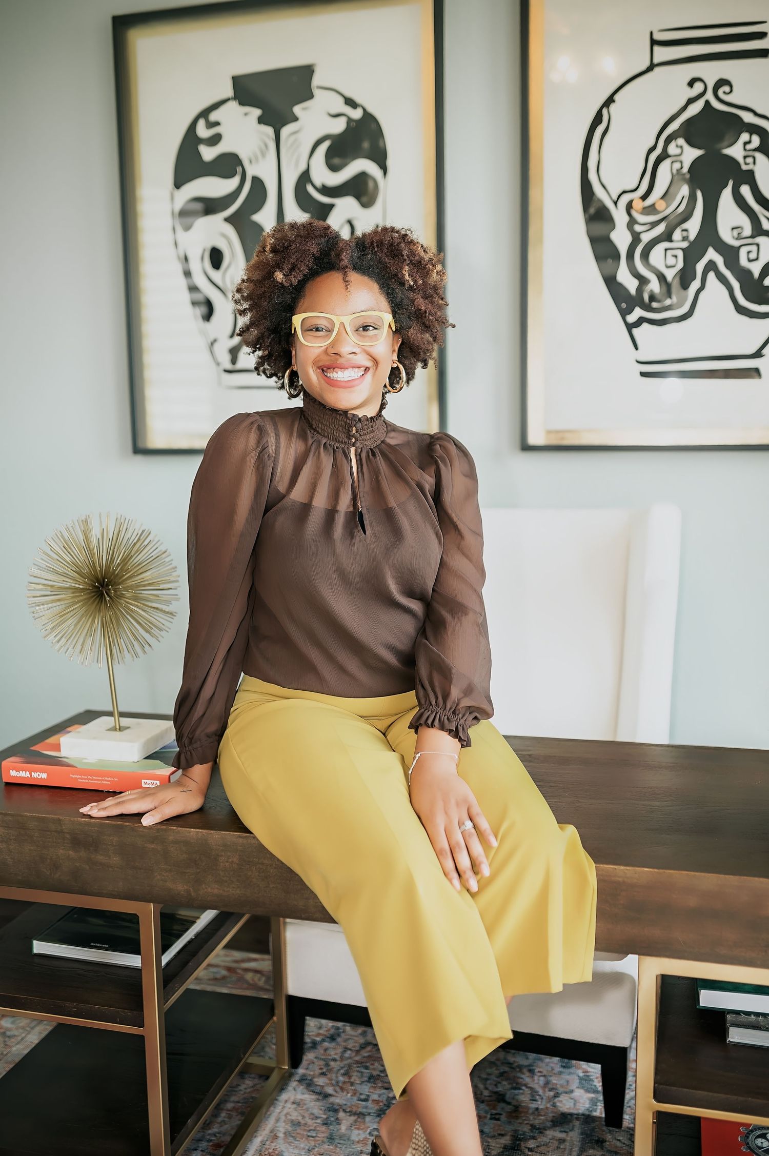 A professional sits on a wooden desk wearing a brown blouse and yellow pants with modern wall art behind them.