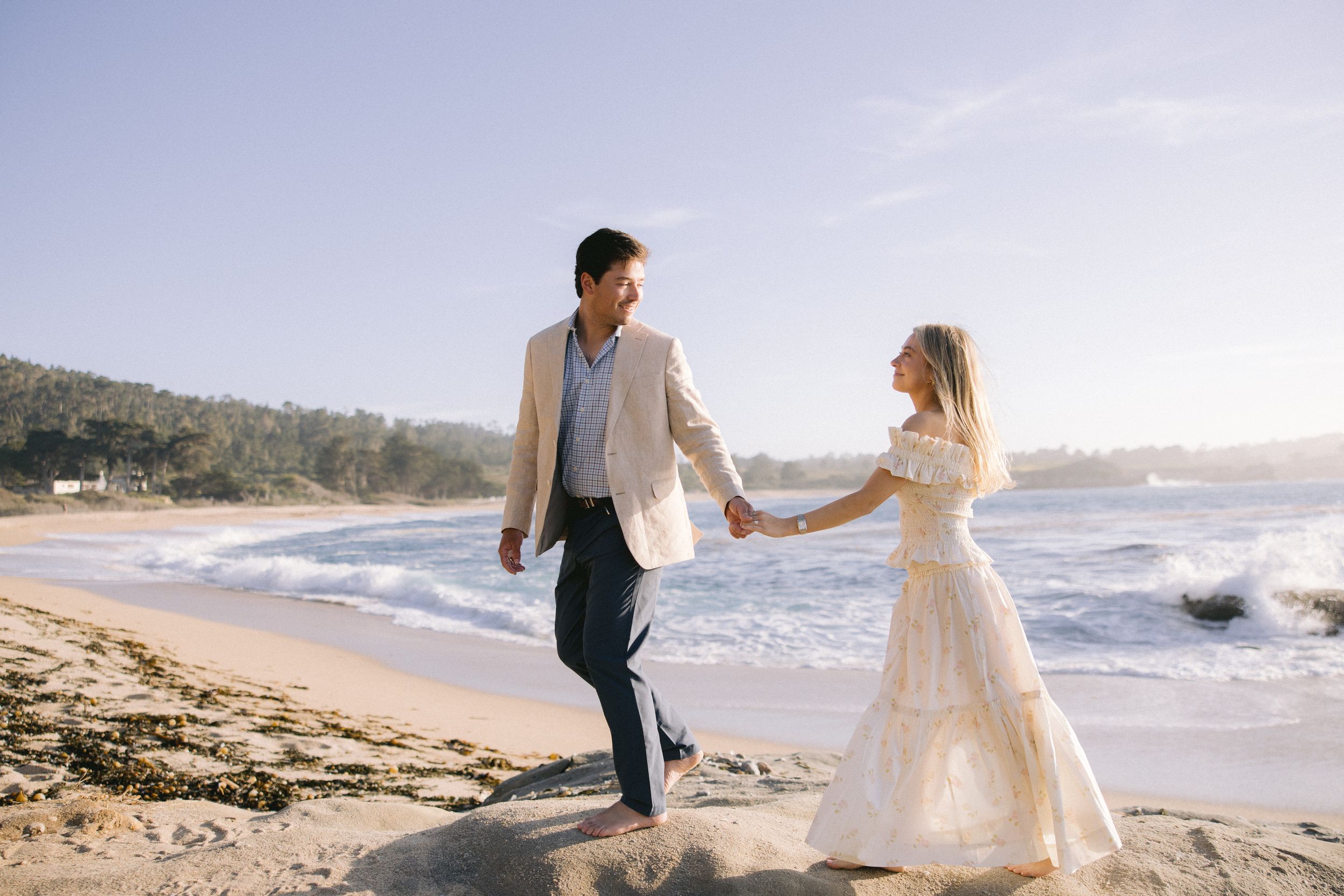 Two people hold hands while walking along a sunny beach shore in formal wedding attire.
