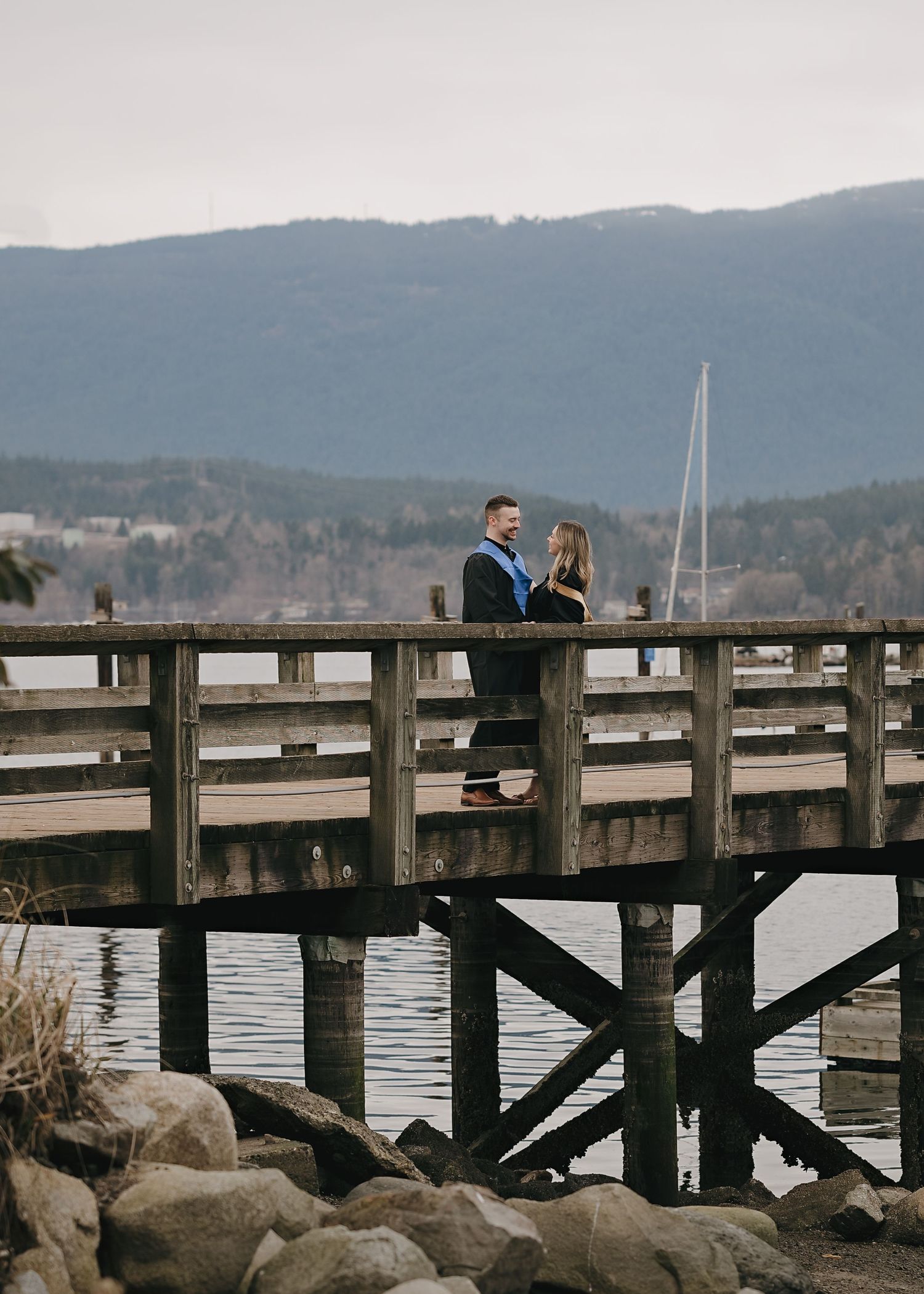 A couple shares an intimate moment on a wooden pier overlooking mountains and water with a sailboat visible in the distance.