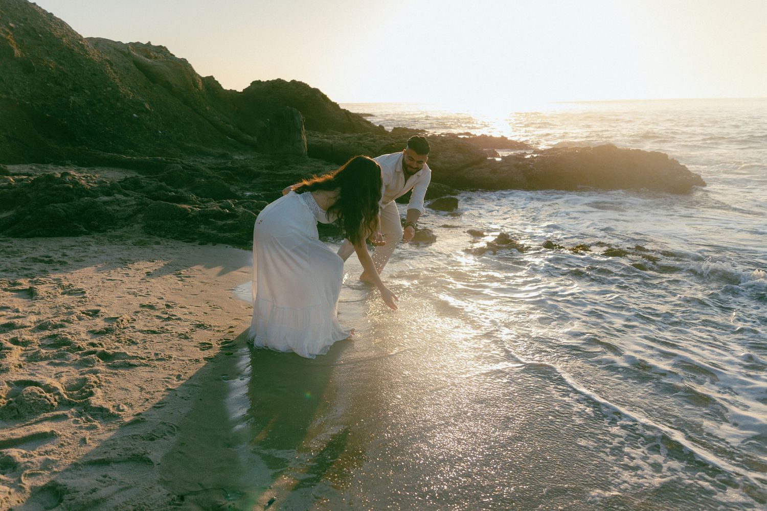 Two people in white dresses stand on rocky beach during golden sunset as waves crash against the shore.