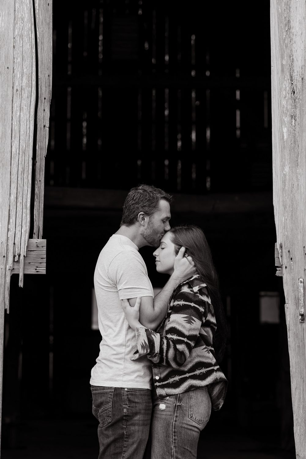 A romantic black and white photo of a couple sharing a tender moment in a rustic barn doorway.