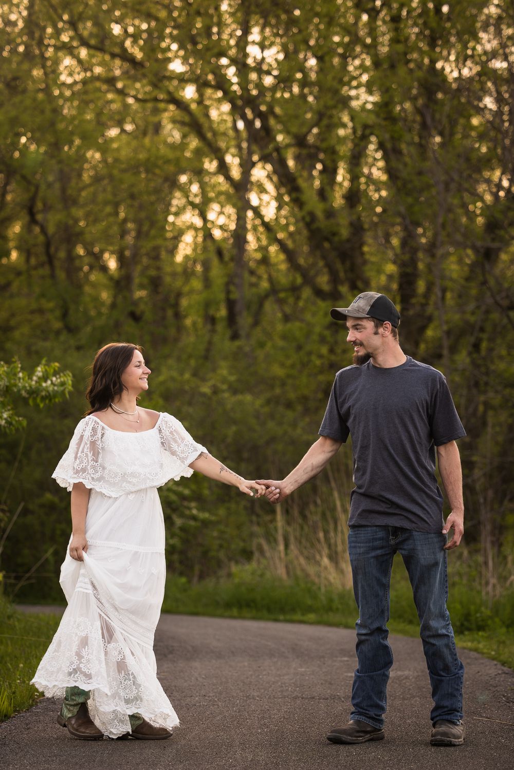 Couple holding hands on path through woods, with woman wearing flowing white dress.