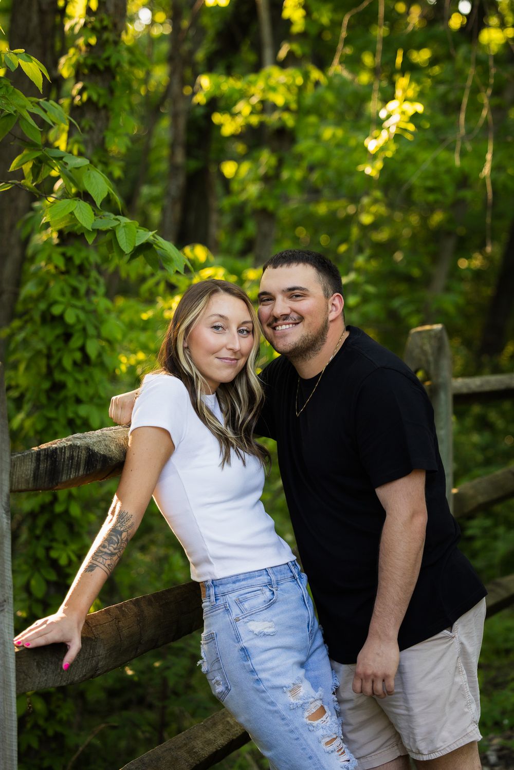 Romantic couple posing by wooden fence in sunlit forest setting during outdoor photoshoot.