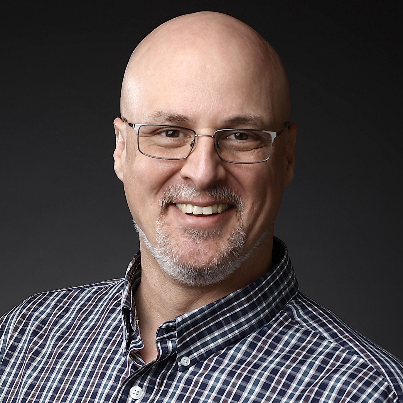 Professional headshot of a smiling person wearing glasses and a plaid shirt against a dark background.