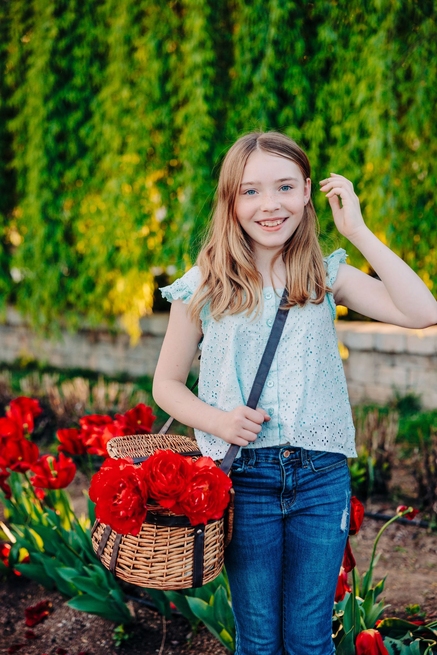 A young person wearing a blue top and jeans poses in a garden with a basket among vibrant red tulips.