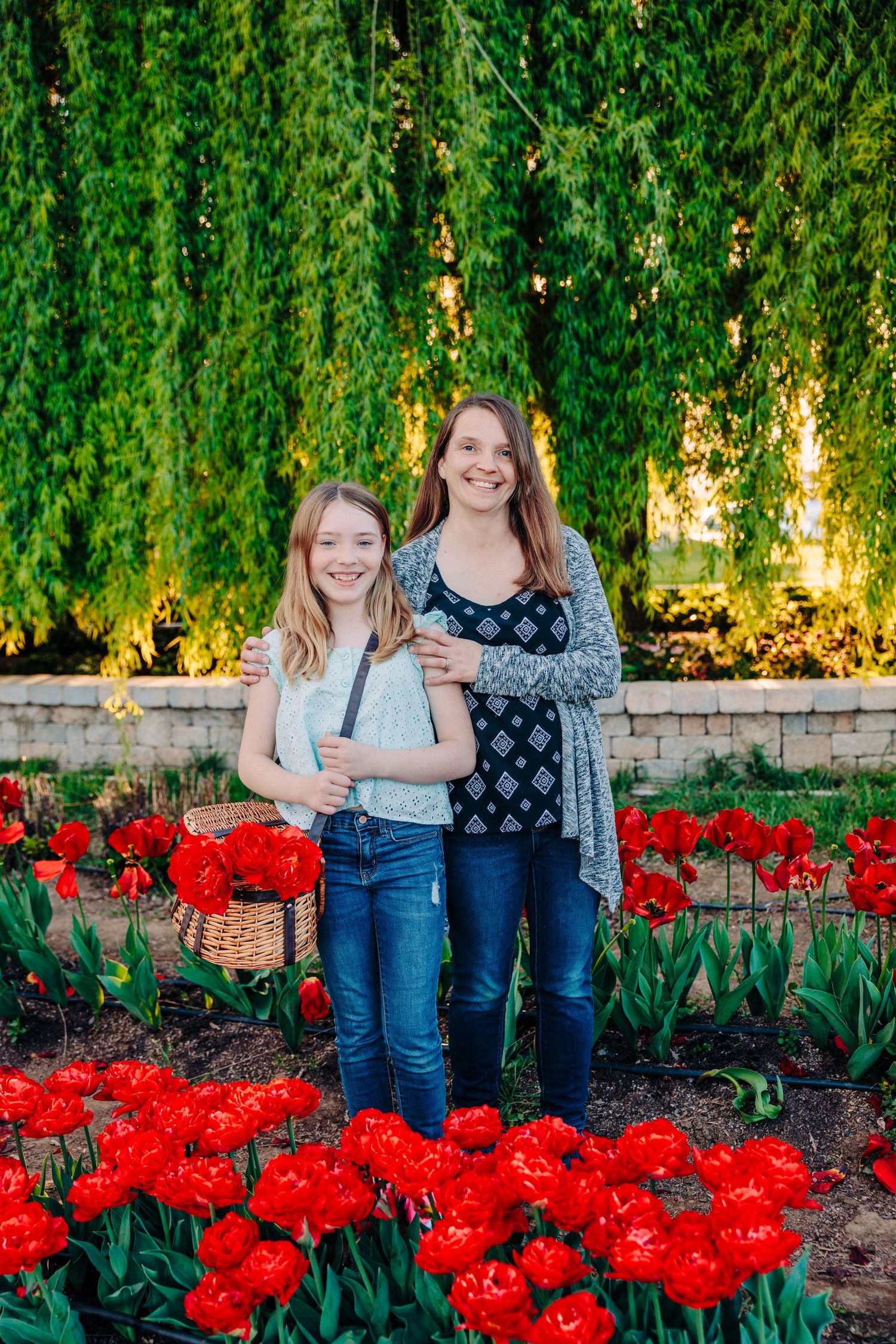 Two people stand together among red tulips in a garden with a cascading willow tree backdrop.