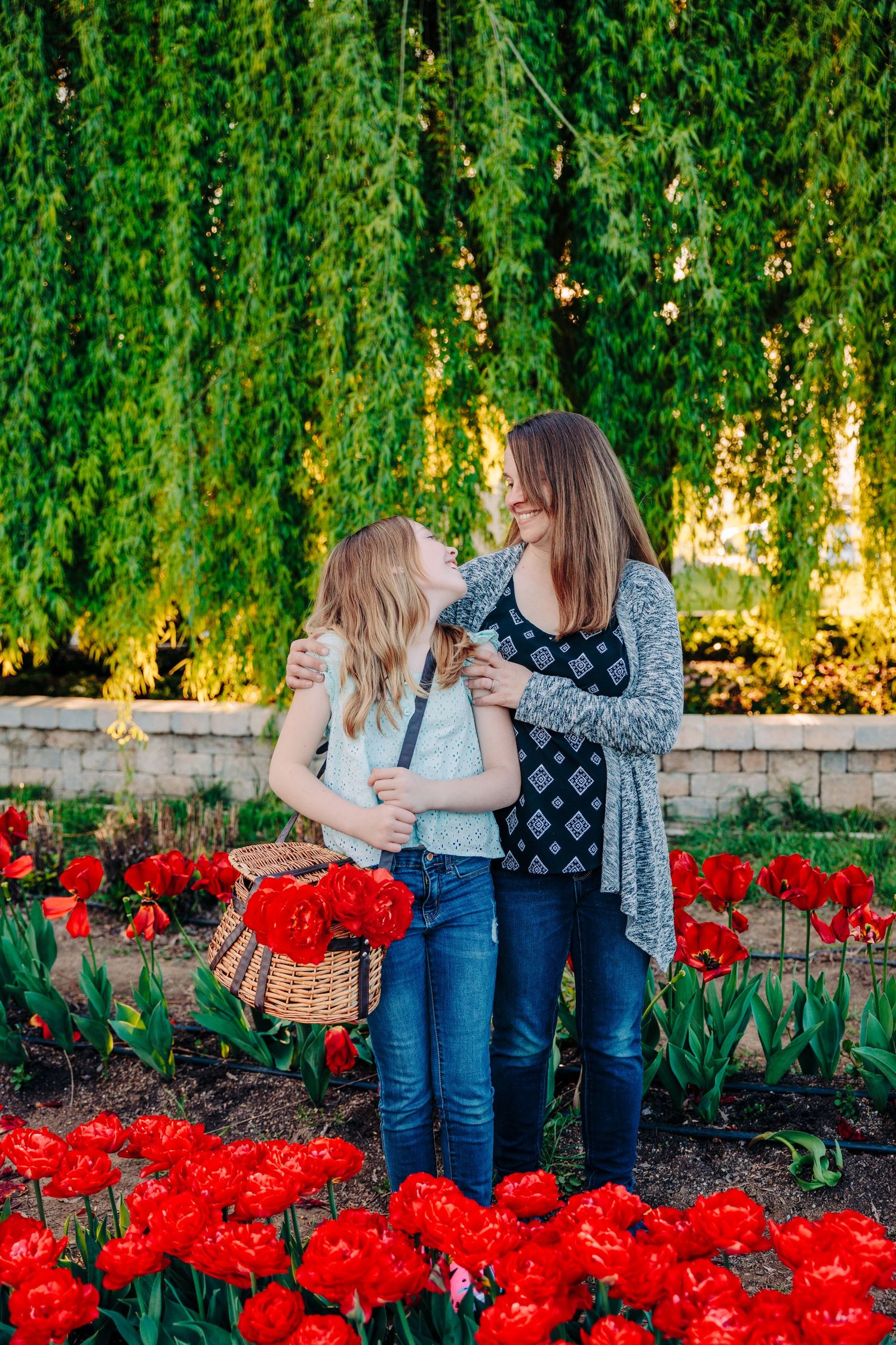 Two people share a joyful moment among vibrant red tulips and cascading willow branches in a garden setting.
