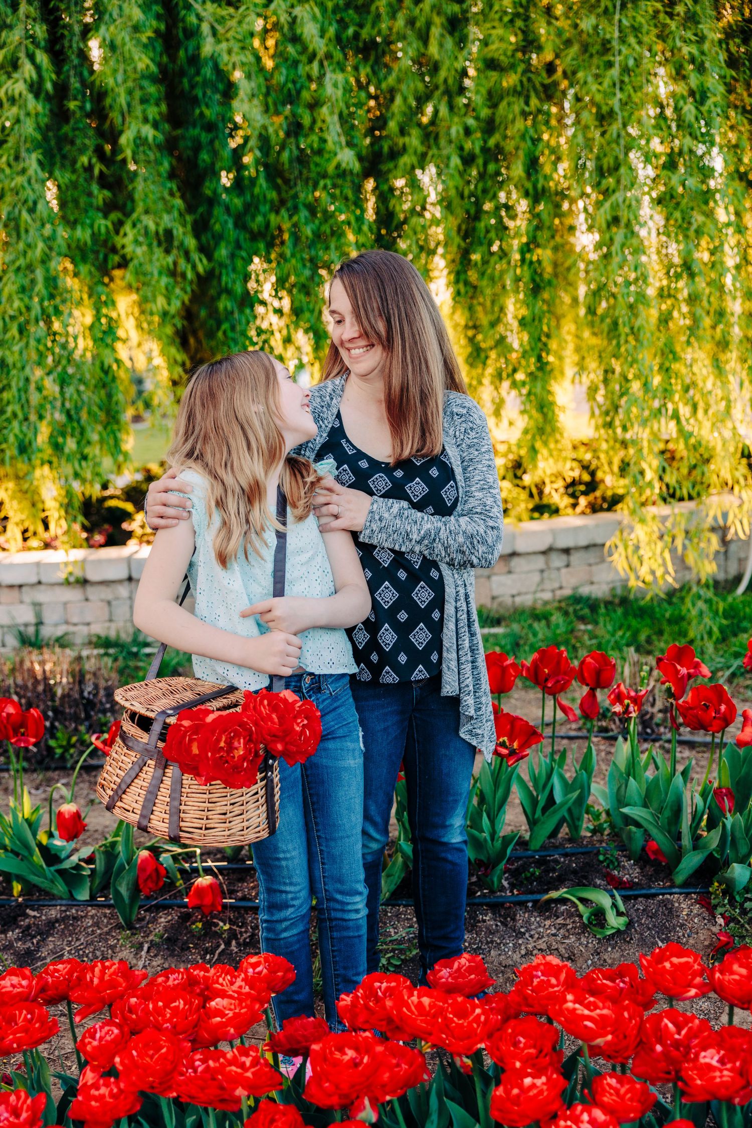 Two people share a joyful moment among vibrant red tulips and drooping willow branches in a garden setting.