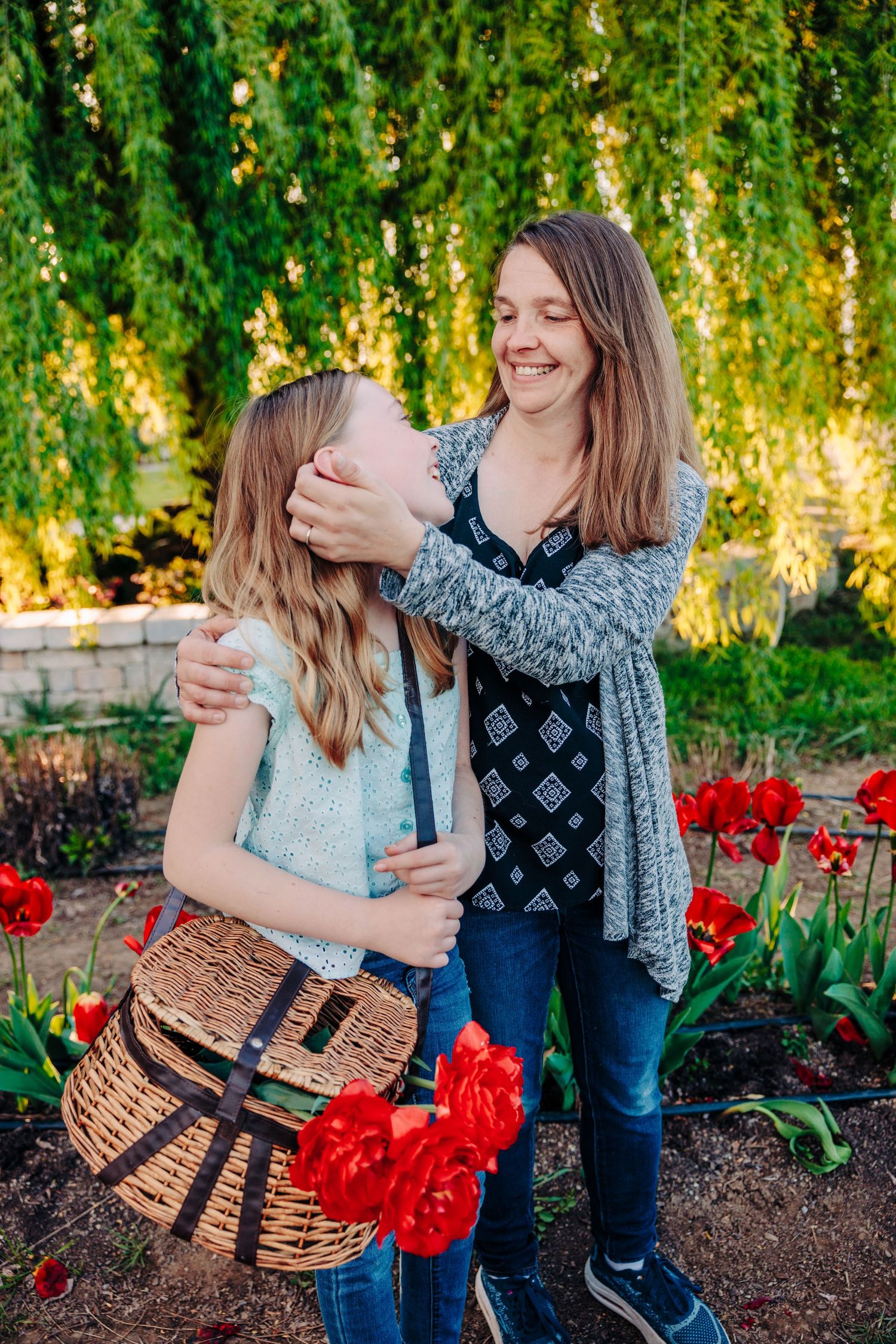 Two people embrace while standing in a garden with bright red tulips and drooping willow branches at sunset.