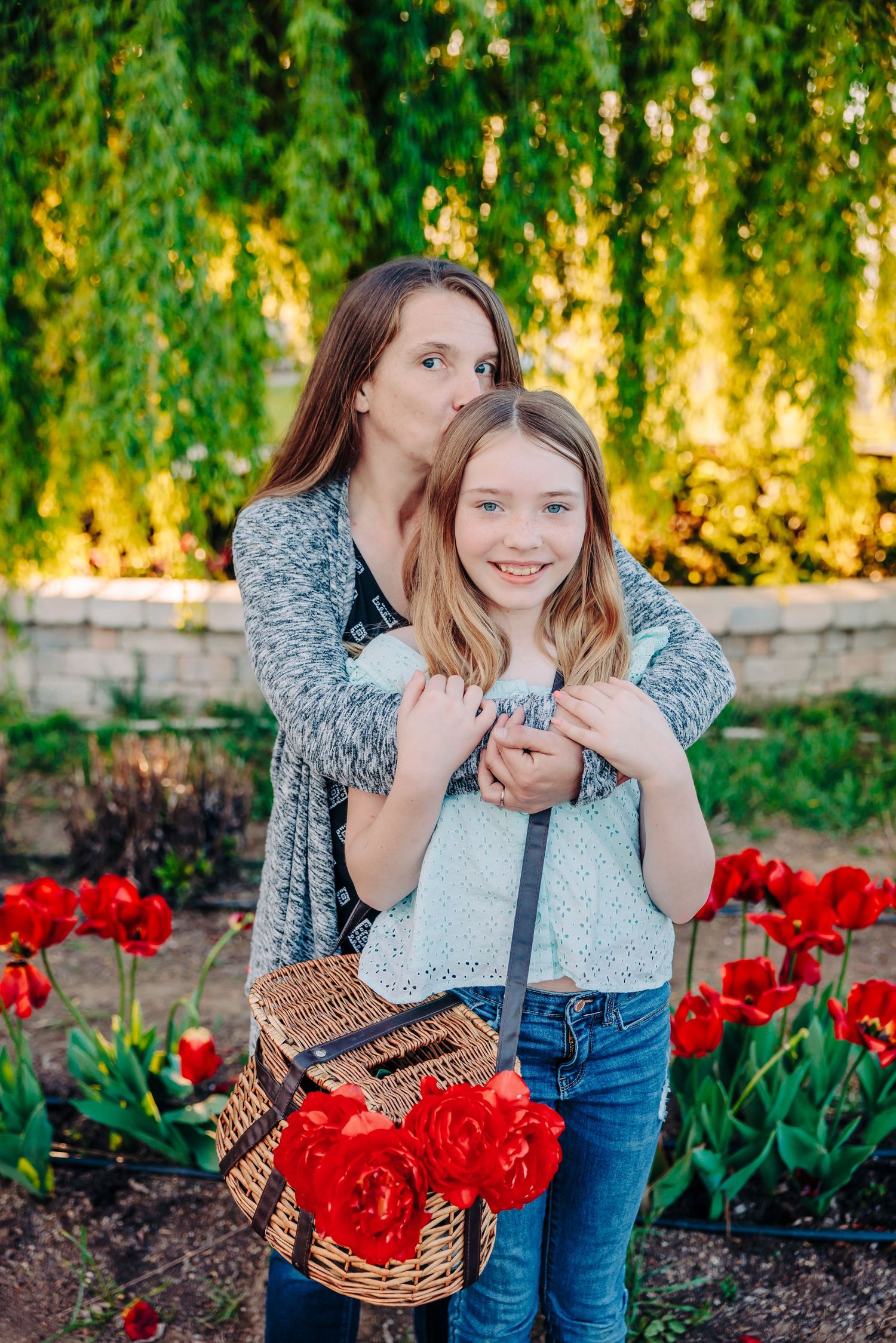 Two people pose in a garden with vibrant red tulips and a wicker picnic basket near a weeping willow tree.