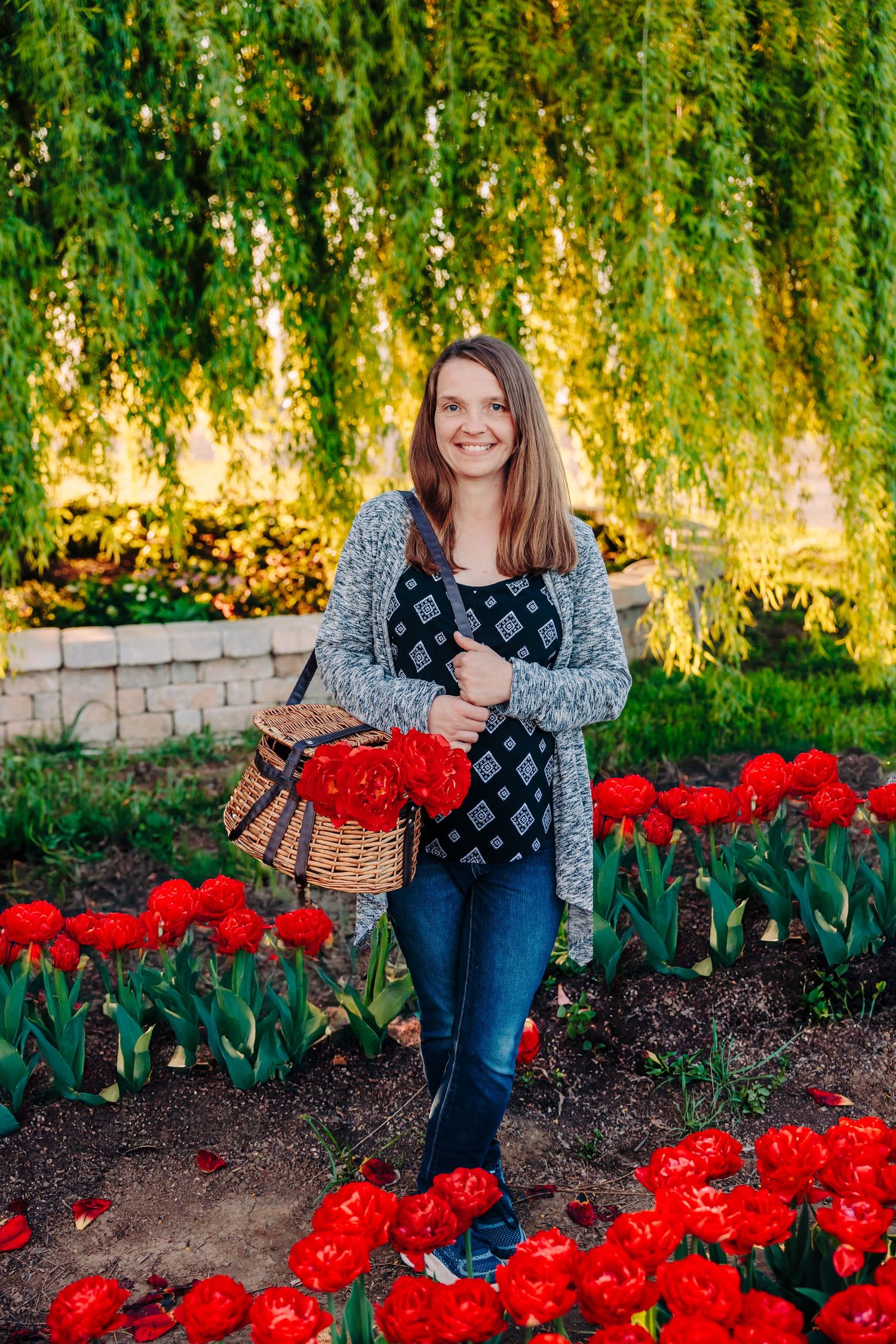 Person in gray cardigan and jeans stands amid blooming red tulips with woven basket.