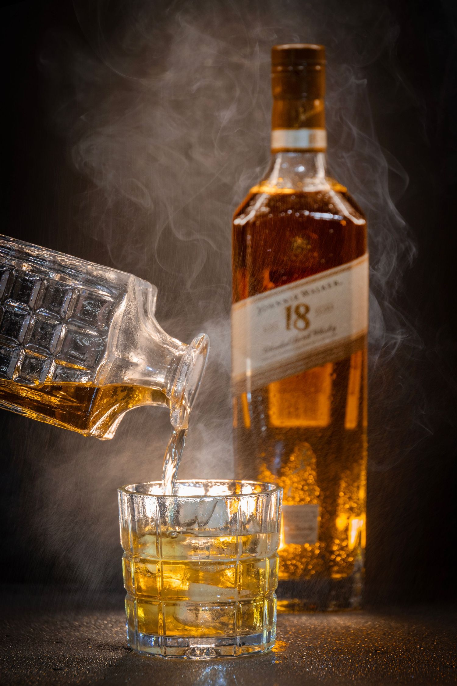 Whisky being poured into a glass with ice from a decanter, beside a bottle of Johnnie Walker Gold Label Reserve with misty backlight.