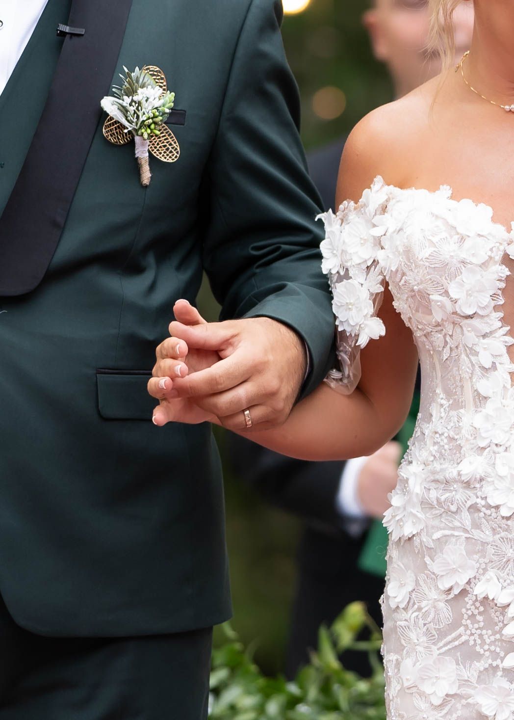 A close-up of wedding attire showing a dark green suit jacket with floral boutonniere next to a white lace bridal gown.