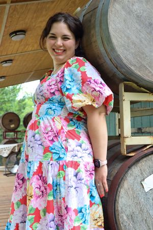 Person wearing colorful floral dress poses by large wooden barrels at a rustic venue.
