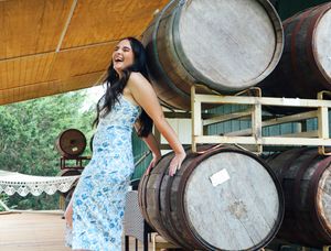 Woman in blue floral dress poses by wooden wine barrels at a winery.