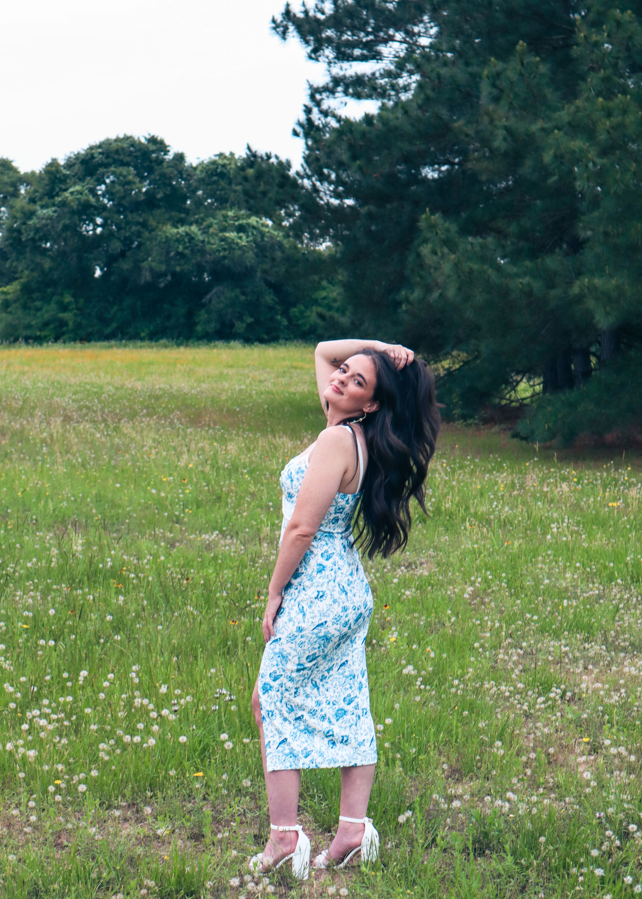 Woman in blue dress poses in meadow with wildflowers and trees.