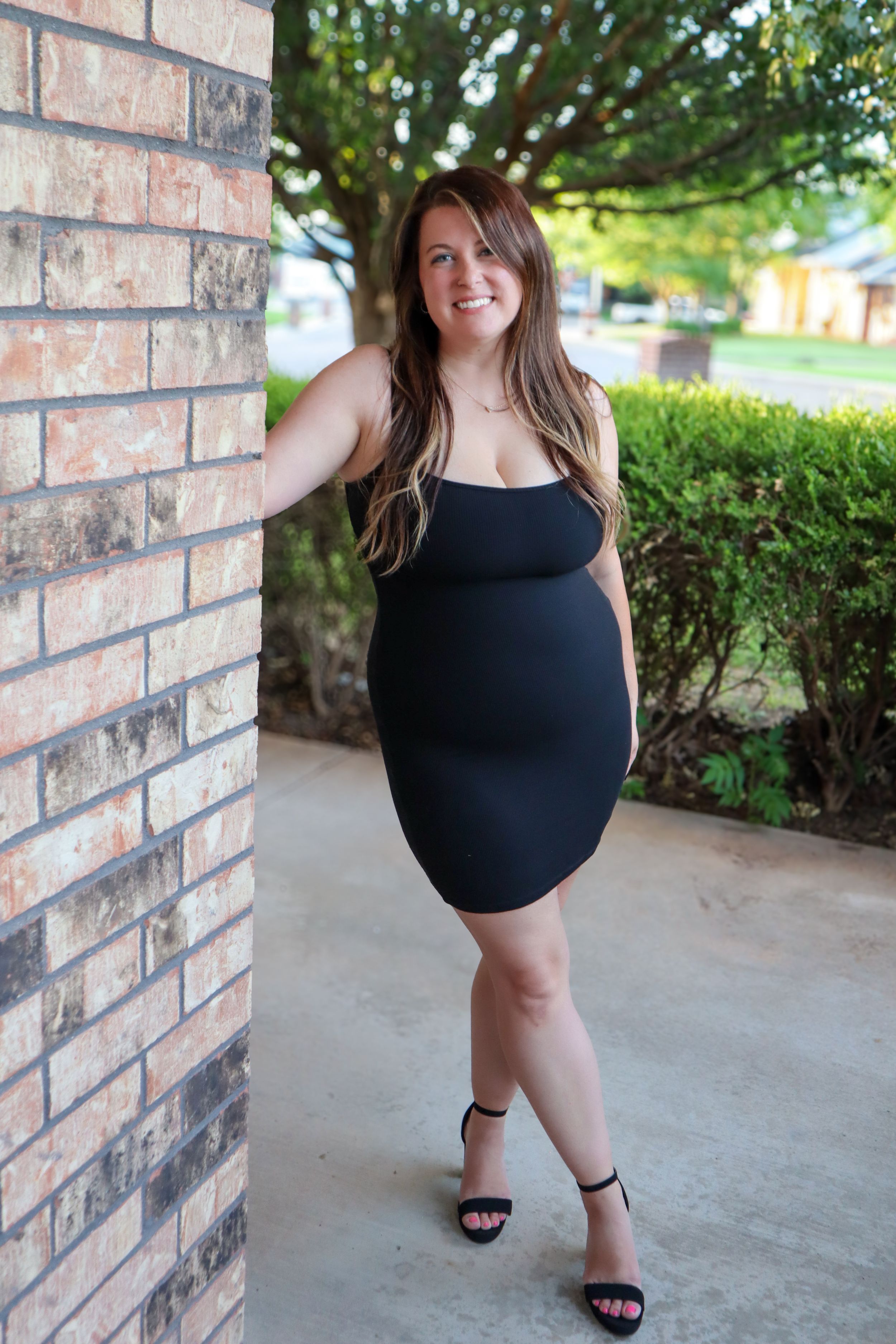 Woman in black dress poses against brick wall outdoors.