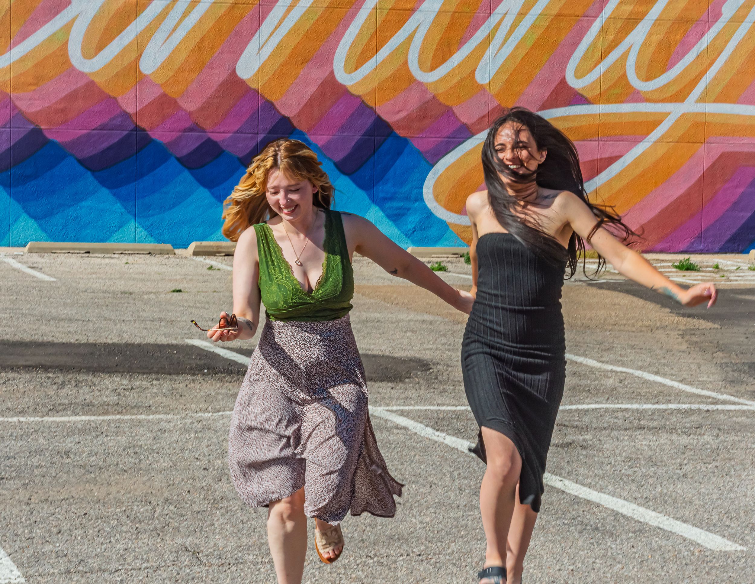 Two friends run and laugh together in front of a colorful geometric mural wall.