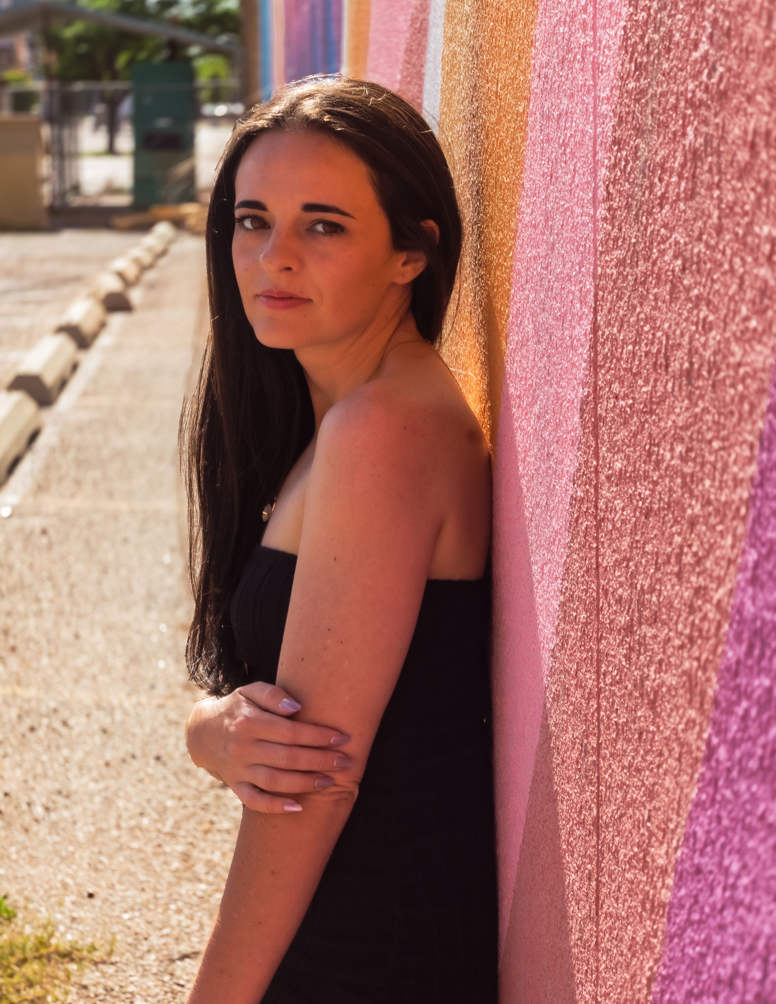 A person in a black top poses against a pink wall in warm sunlight.