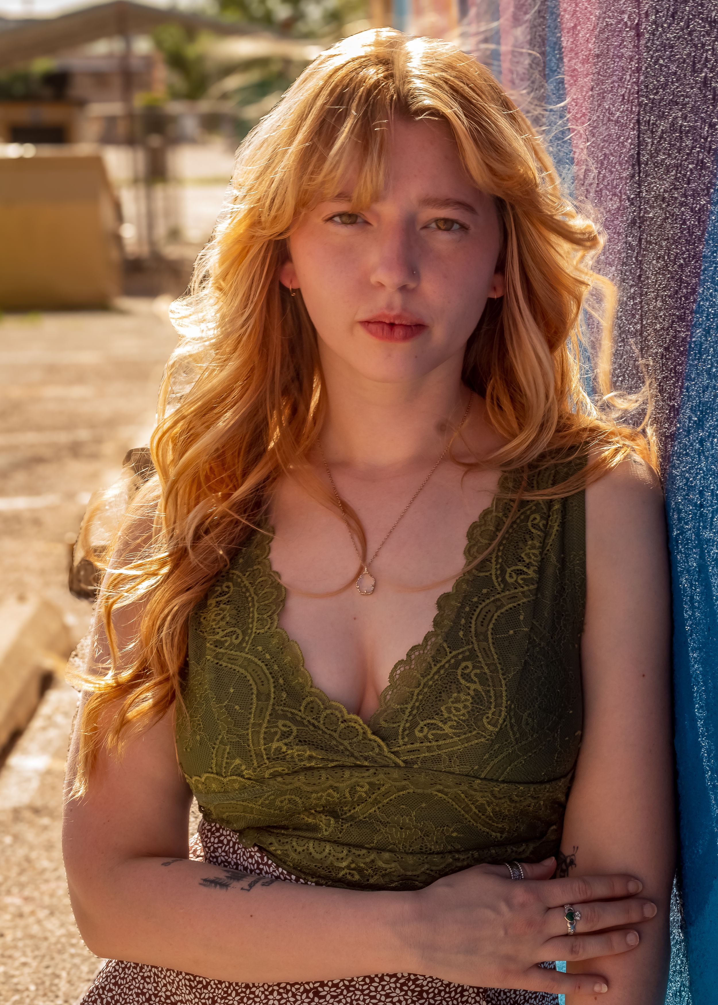 Woman with long wavy golden hair poses in olive green lace top against sunlit wall outdoors.
