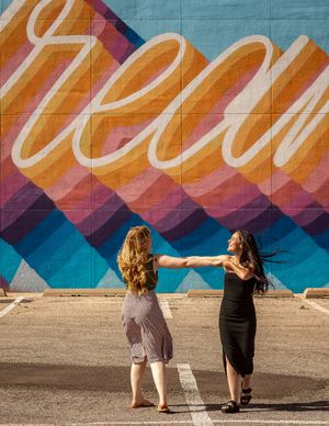 Two friends hold hands and dance in front of a large decorative wall mural with flowing text.