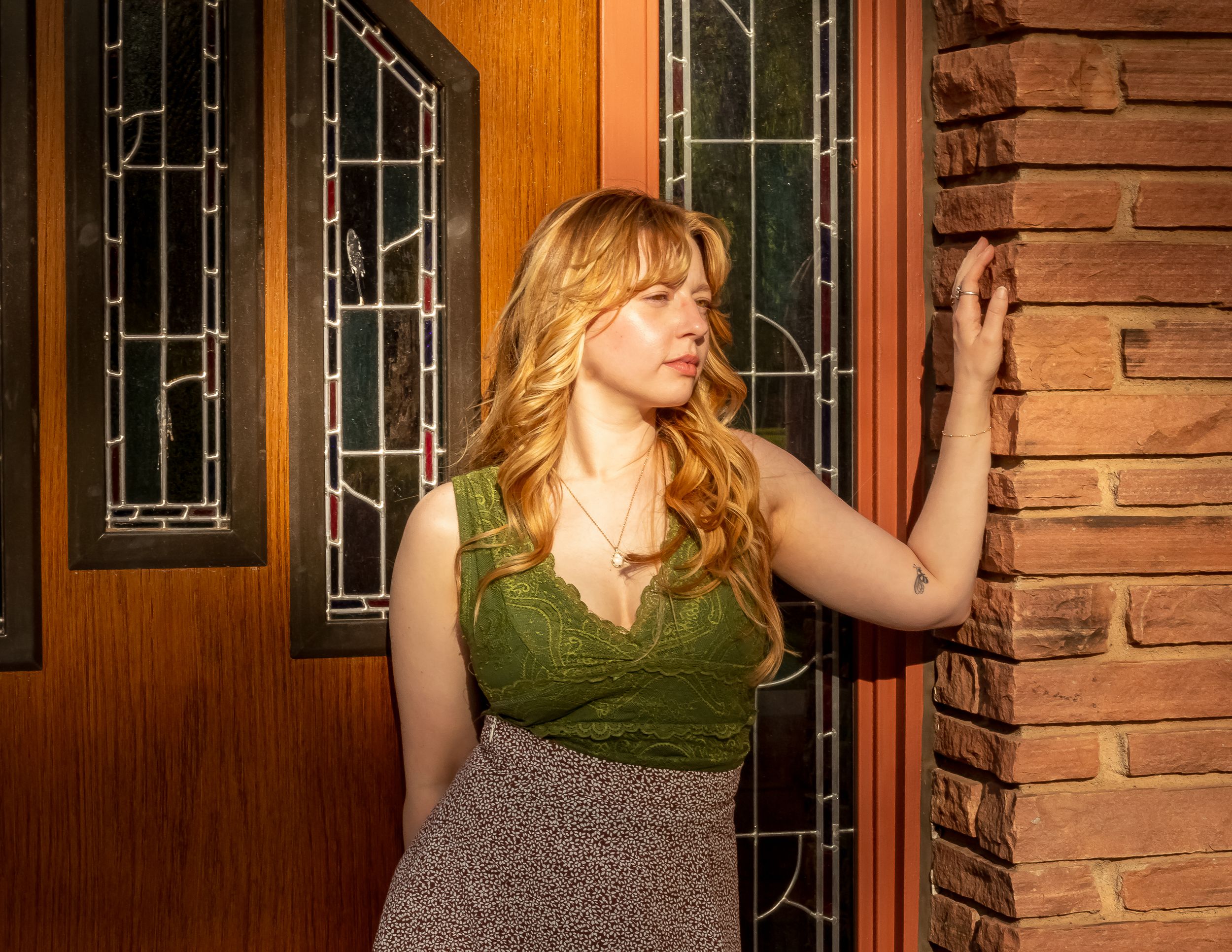 A woman in a green sleeveless top and patterned skirt poses by a wooden door with decorative glass panels.