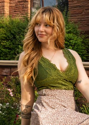 Person wearing green lace top and patterned skirt poses outdoors against brick wall with greenery.