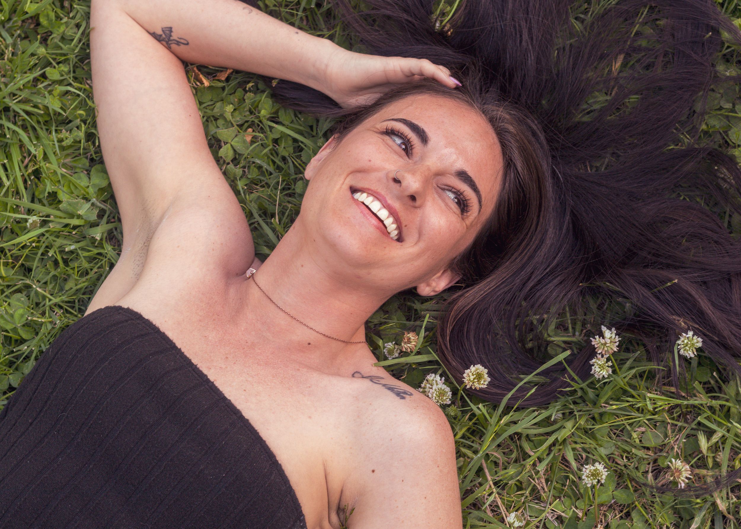A person in black lies in grass with flowers while laughing joyfully at the sky.