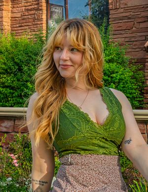 Person in green lace top and floral skirt poses by brick wall with greenery in background.