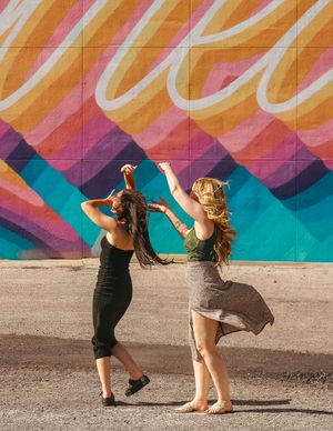 Two women dancing playfully in front of vibrant orange and pink street art mural.