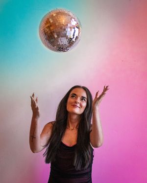 A person tosses and catches a silver disco ball against a pink and turquoise colored backdrop in a sequence of movements.