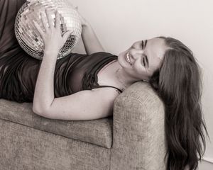 A person lies on a couch holding a disco ball while wearing a dark top in a black and white photo.