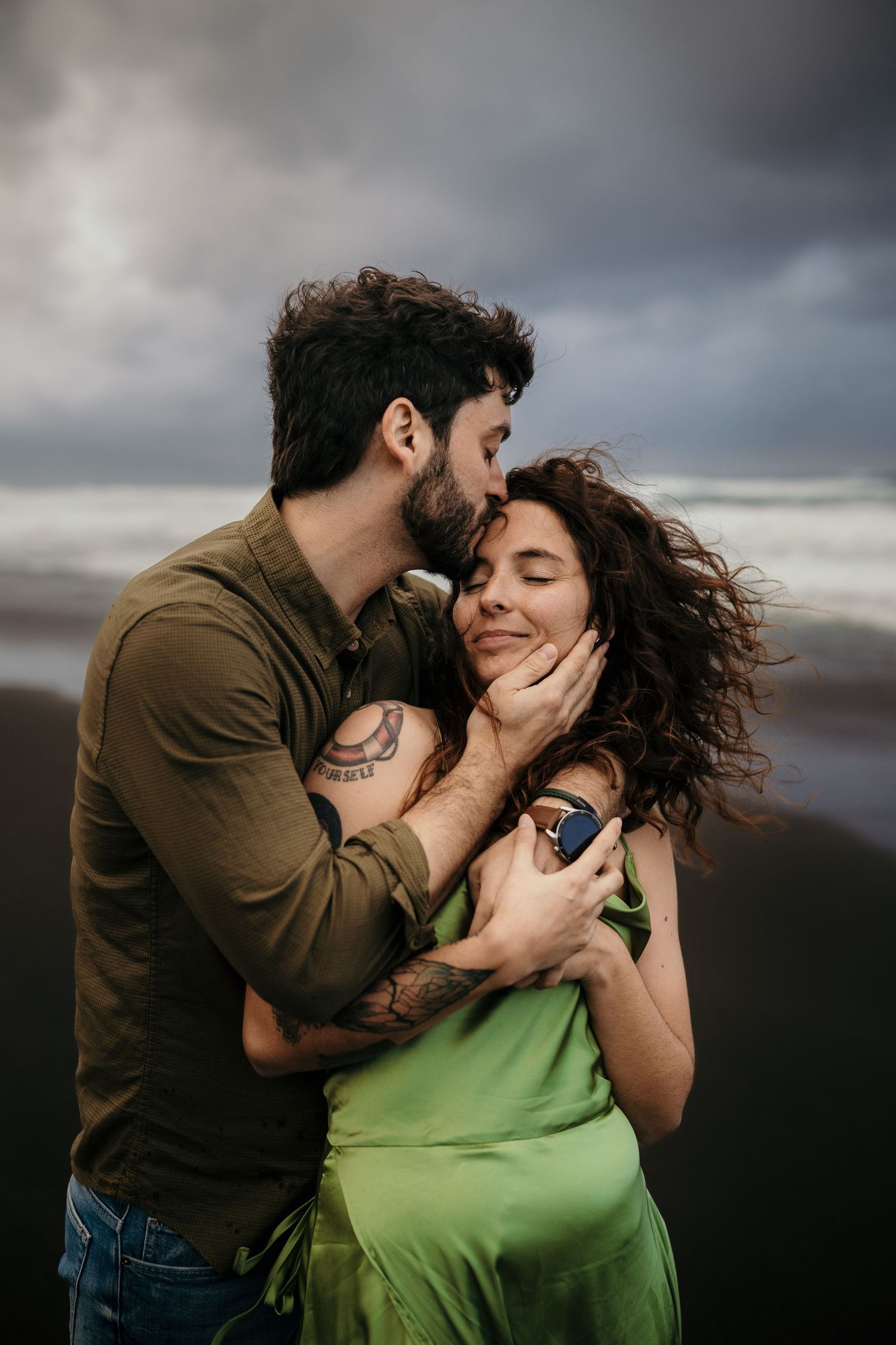 Romantic couple embraces tenderly against moody sky backdrop at beach as wind tousles her green dress and long curly hair.