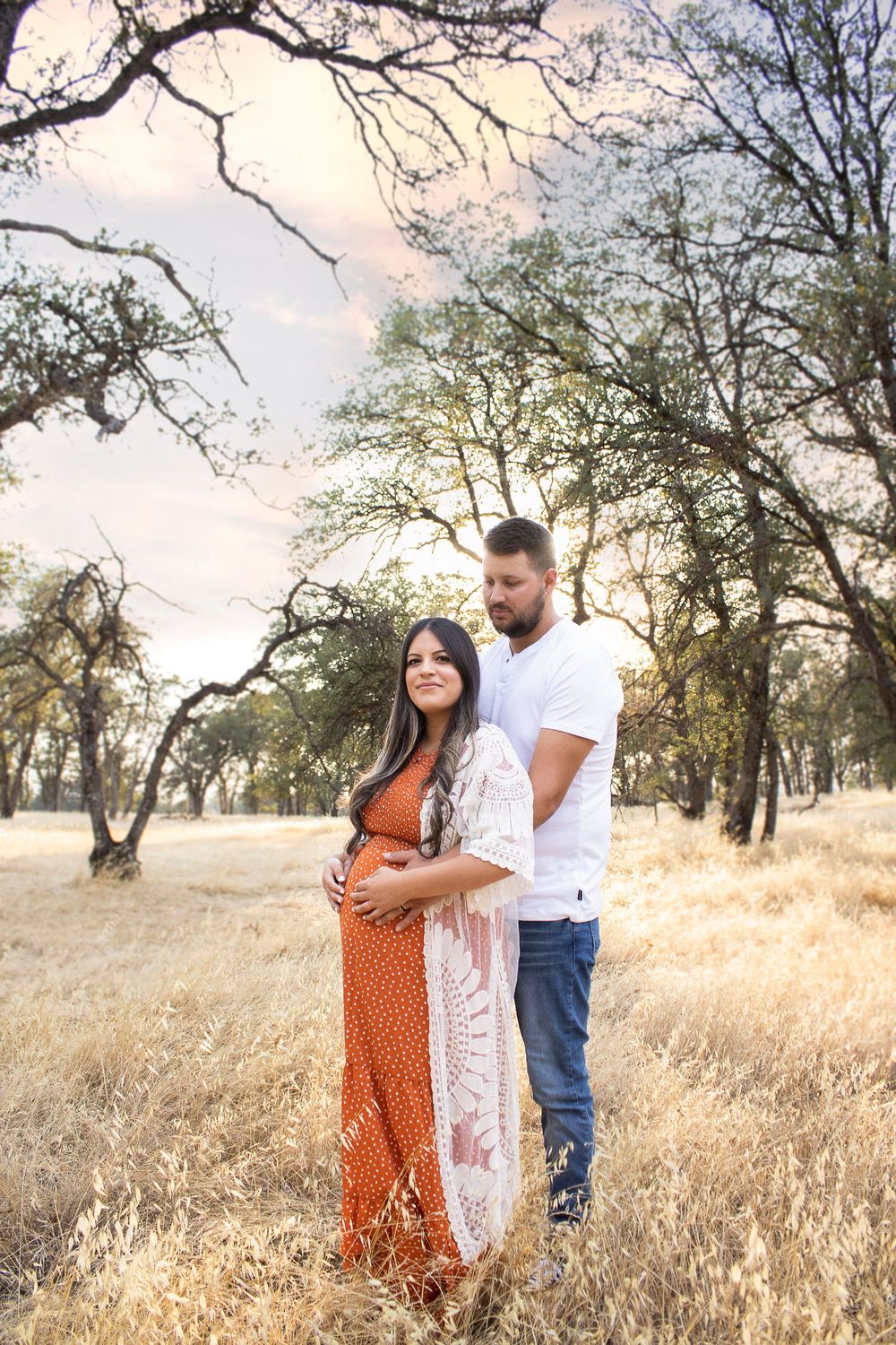 A sunset maternity session in a golden field with the couple wearing white and rust-colored outfits.