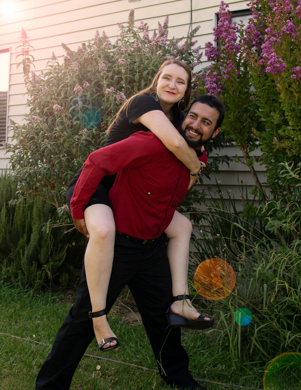 A playful piggyback pose in front of blooming purple flowers and a house exterior.