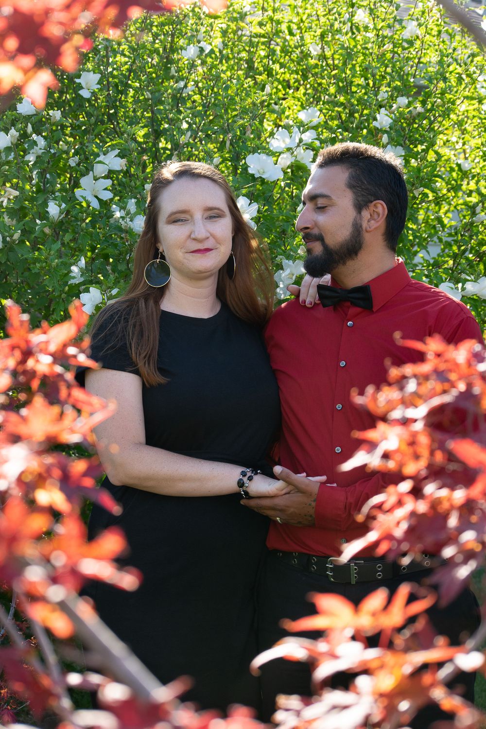 A couple poses among vibrant red and green foliage during an outdoor portrait session.