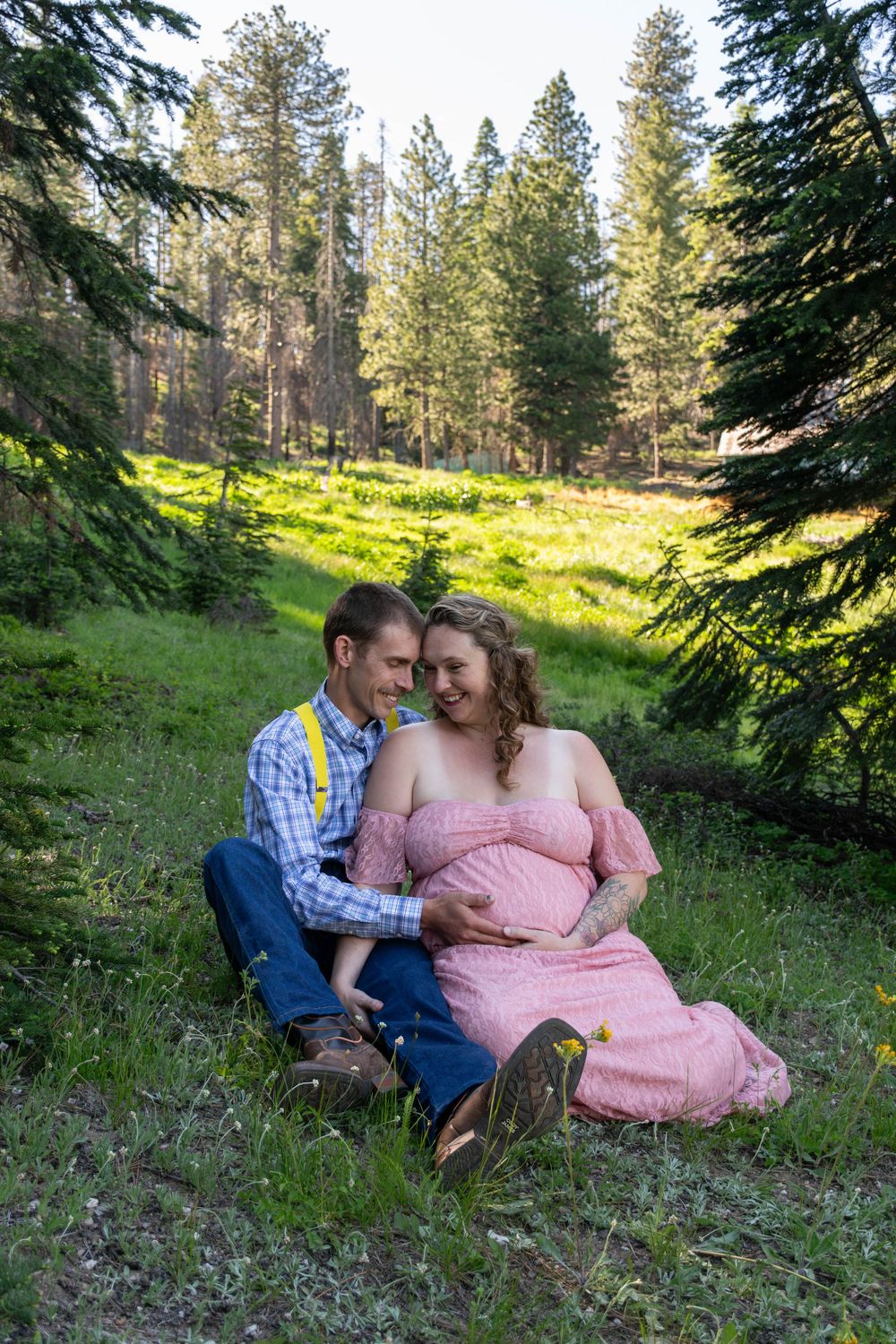 A romantic maternity photo session in a sunlit forest meadow with the couple sitting together on grass.