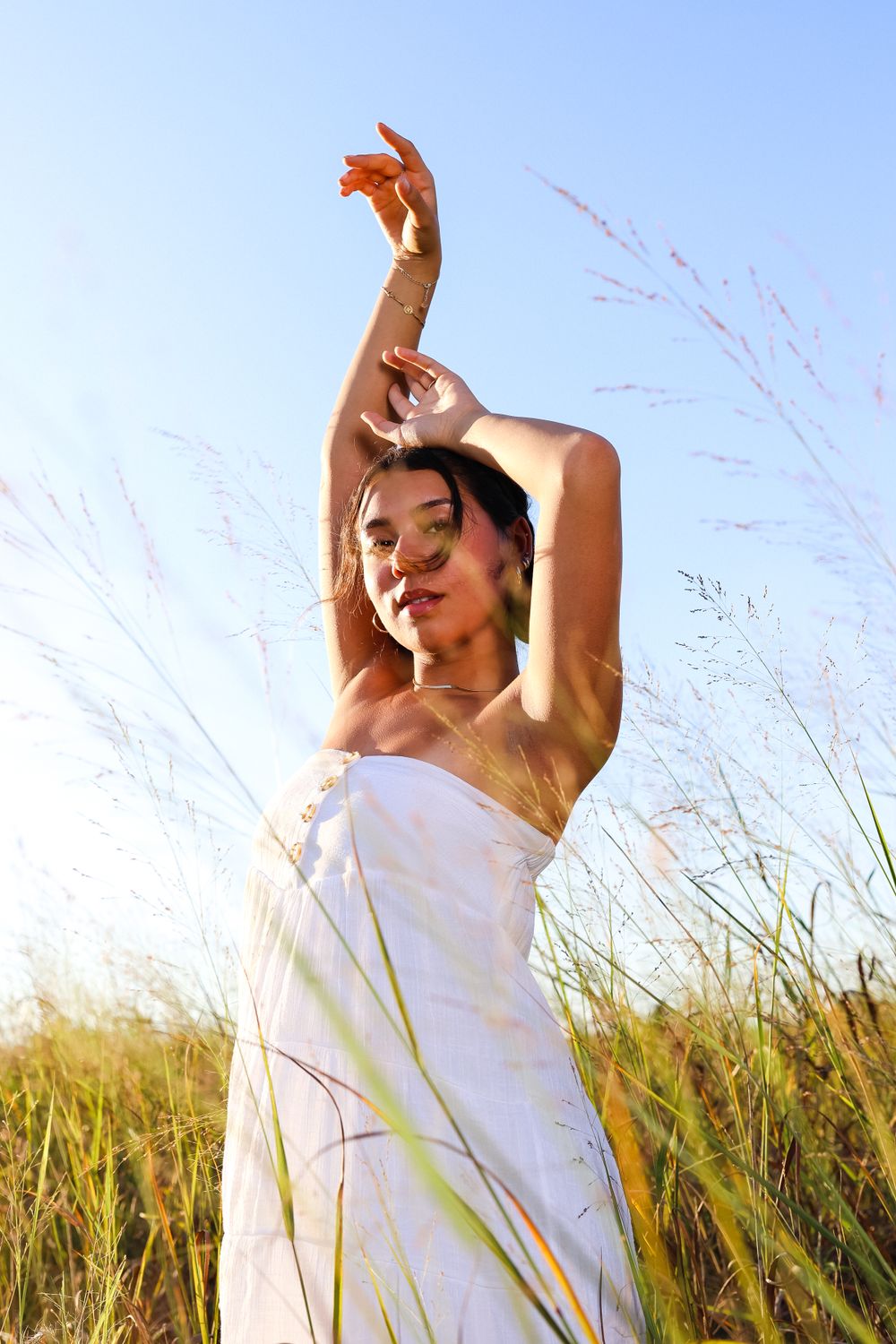 Person in white dress poses gracefully in tall grass against blue sky.
