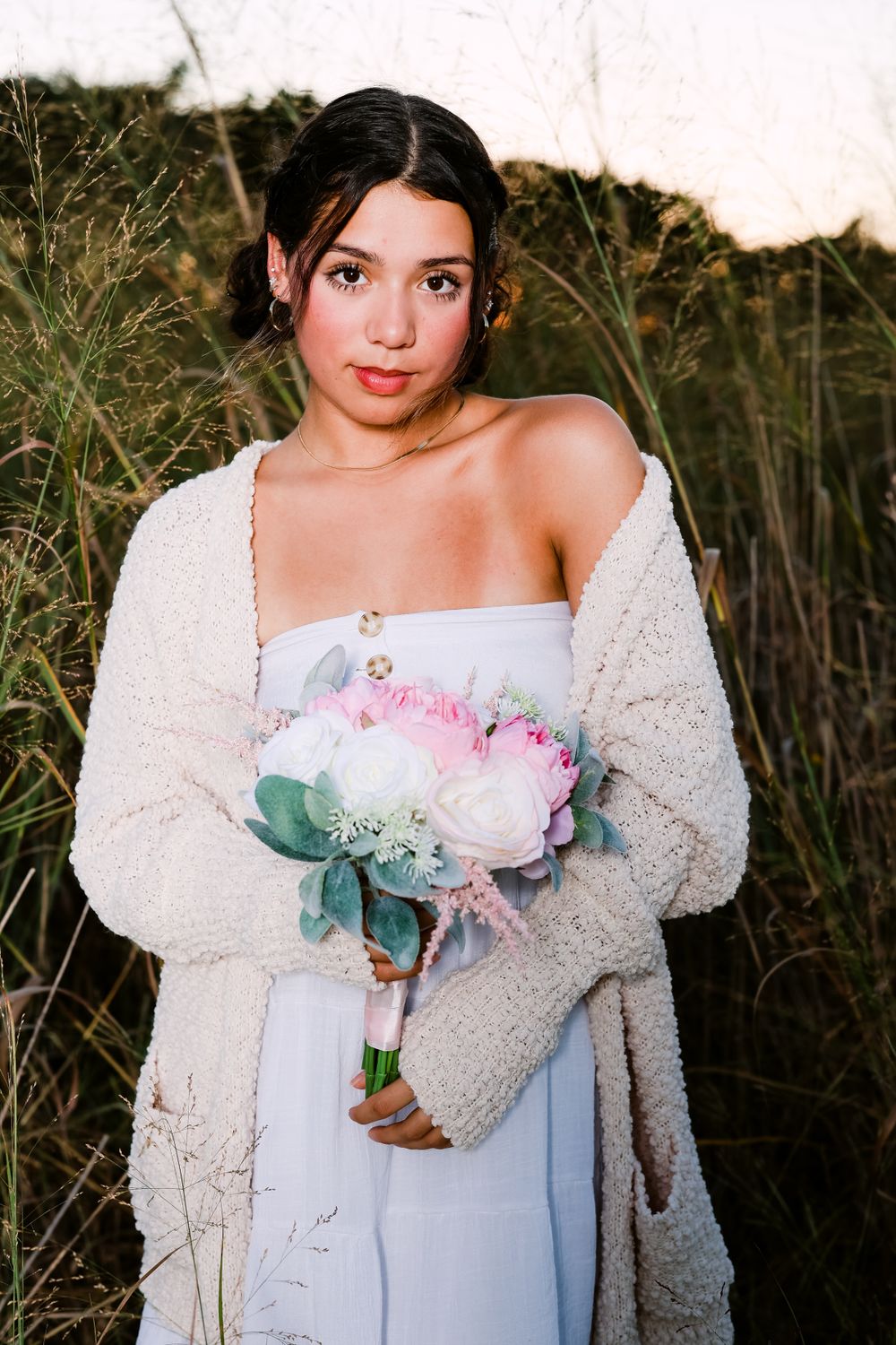 Person in white dress holding pink and white flowers outdoors with fuzzy cardigan.