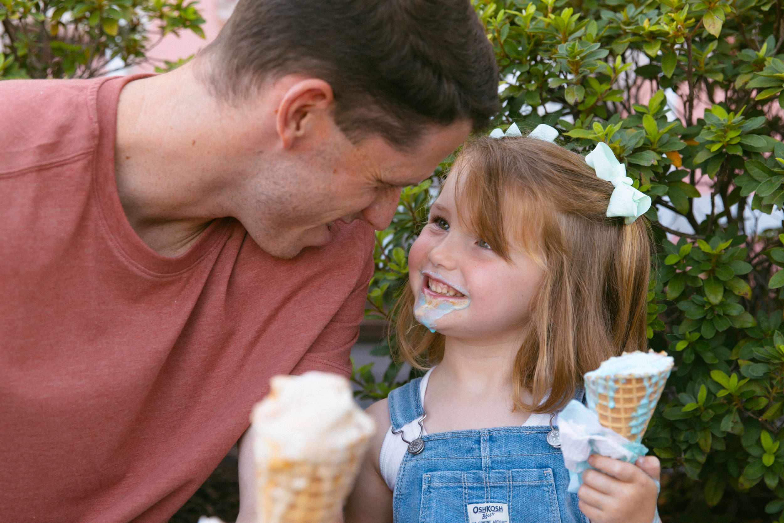 A parent and child sharing ice cream cones together while smiling outdoors.